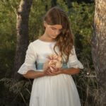 Niña en vestido blanco de comunión sosteniendo flores en un bosque.
