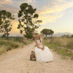 Niña en vestido blanco de comunión sentada en un camino rural al atardecer, rodeada de naturaleza.